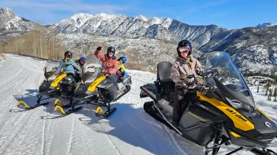Four people riding snowmobiles on a snowy mountain trail with clear blue skies.